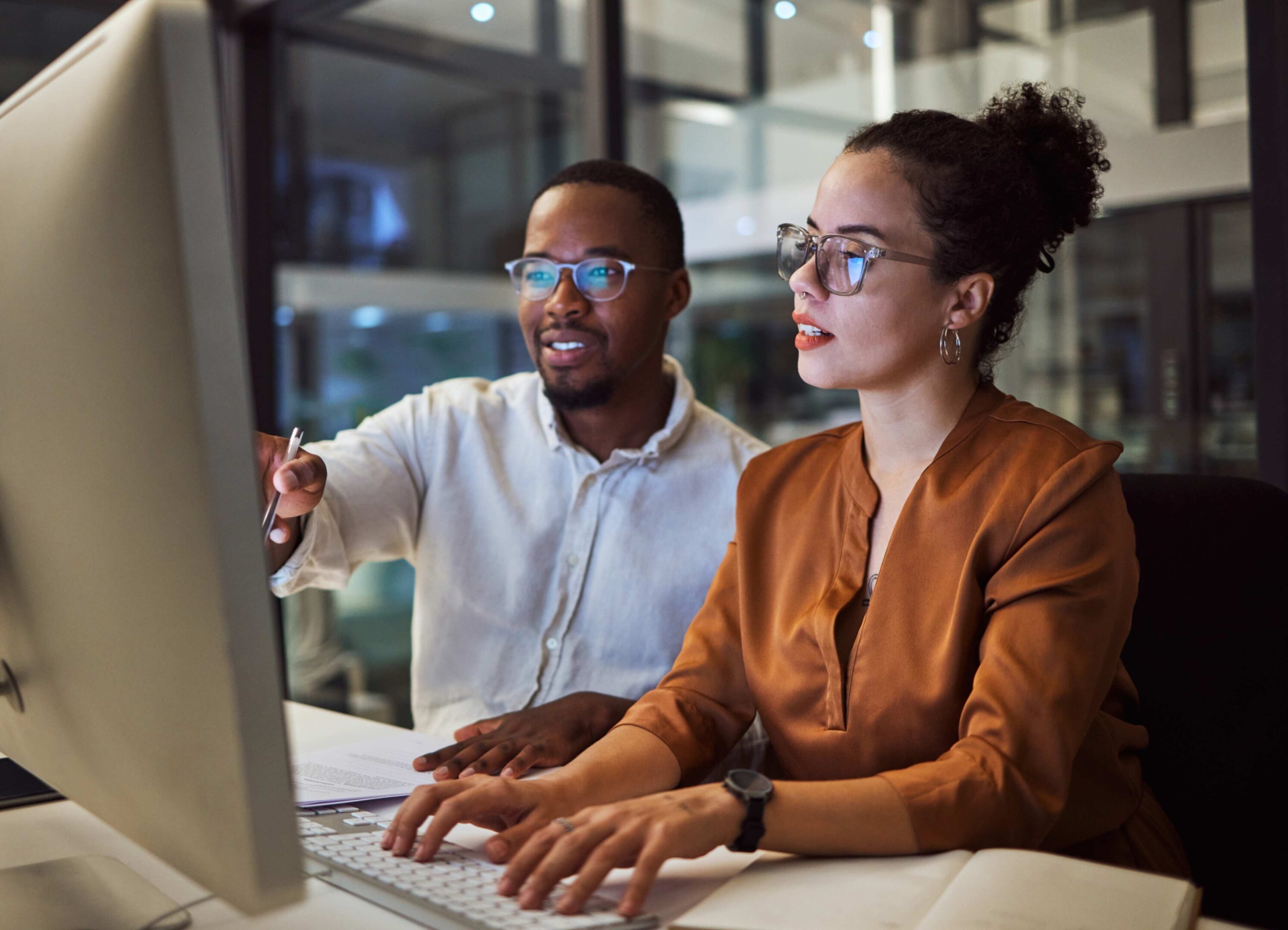 Two people work together on a computer in an office setting. One points at the screen, both wearing glasses.