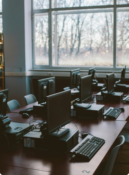 A row of computers on desks in a classroom with large windows showing trees outside.