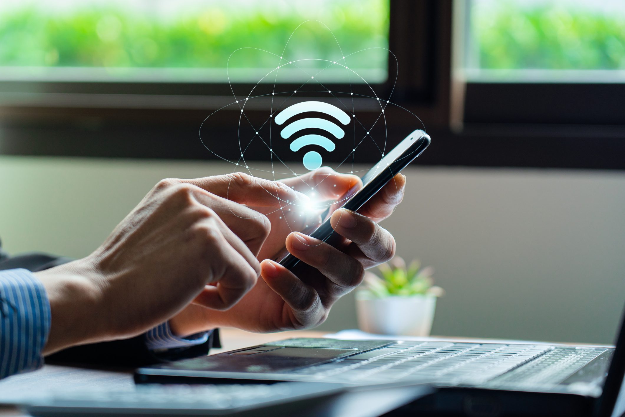 Person using a smartphone with a holographic Wi-Fi symbol above it, near a laptop and a small plant, in front of a window.