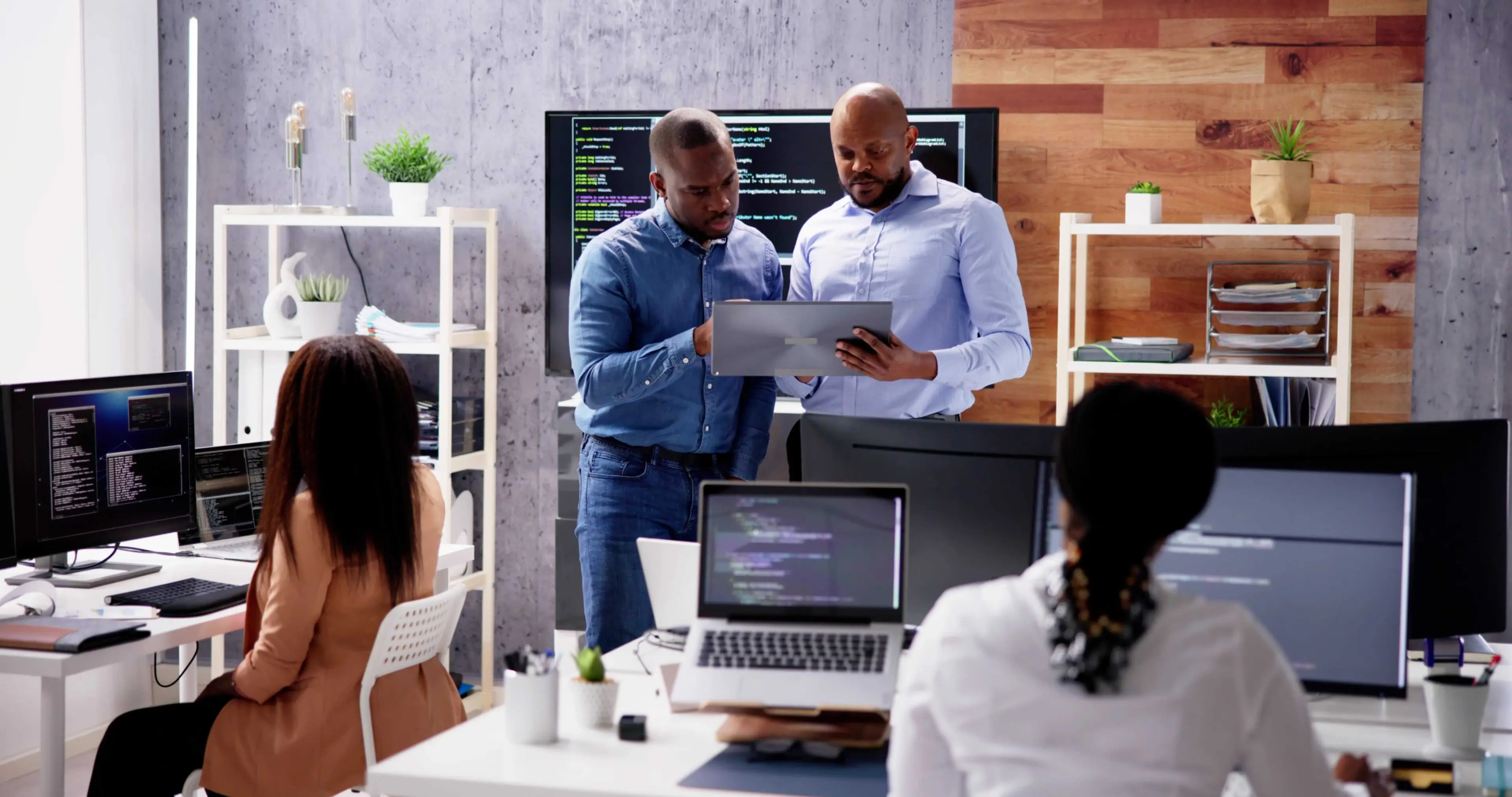 Two men review information on a tablet while two women work at computers in a modern office with monitors displaying code.