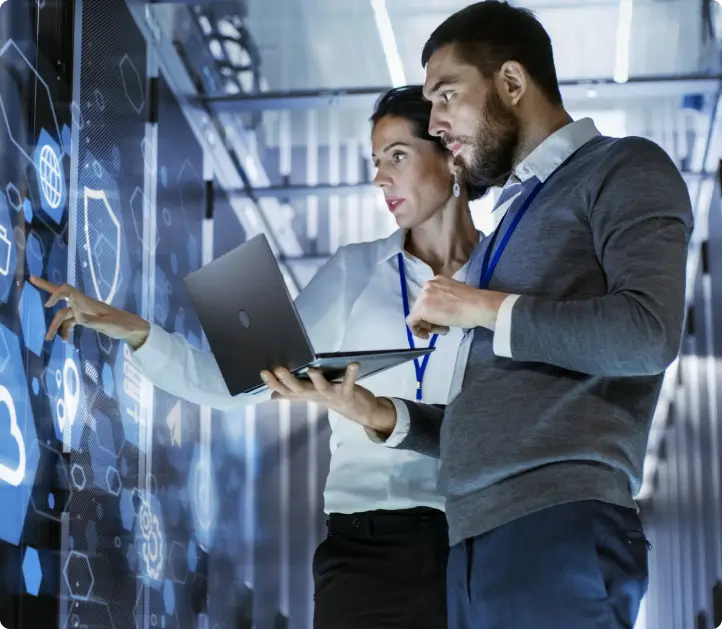 Two people in business attire examine data on a large digital screen in a server room. One holds a laptop while the other points to icons on the screen.