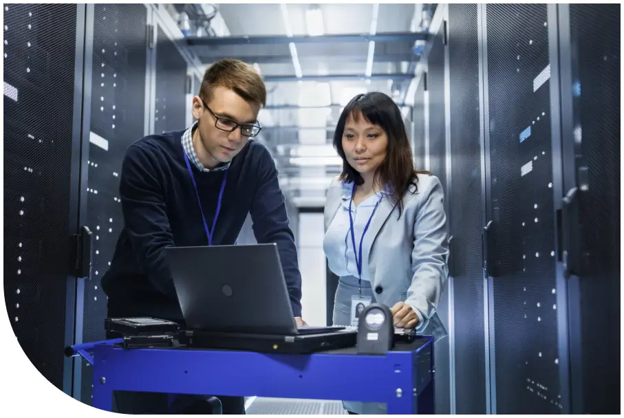 Two people with ID badges stand in a server room, looking at a laptop on a wheeled cart, surrounded by server racks.